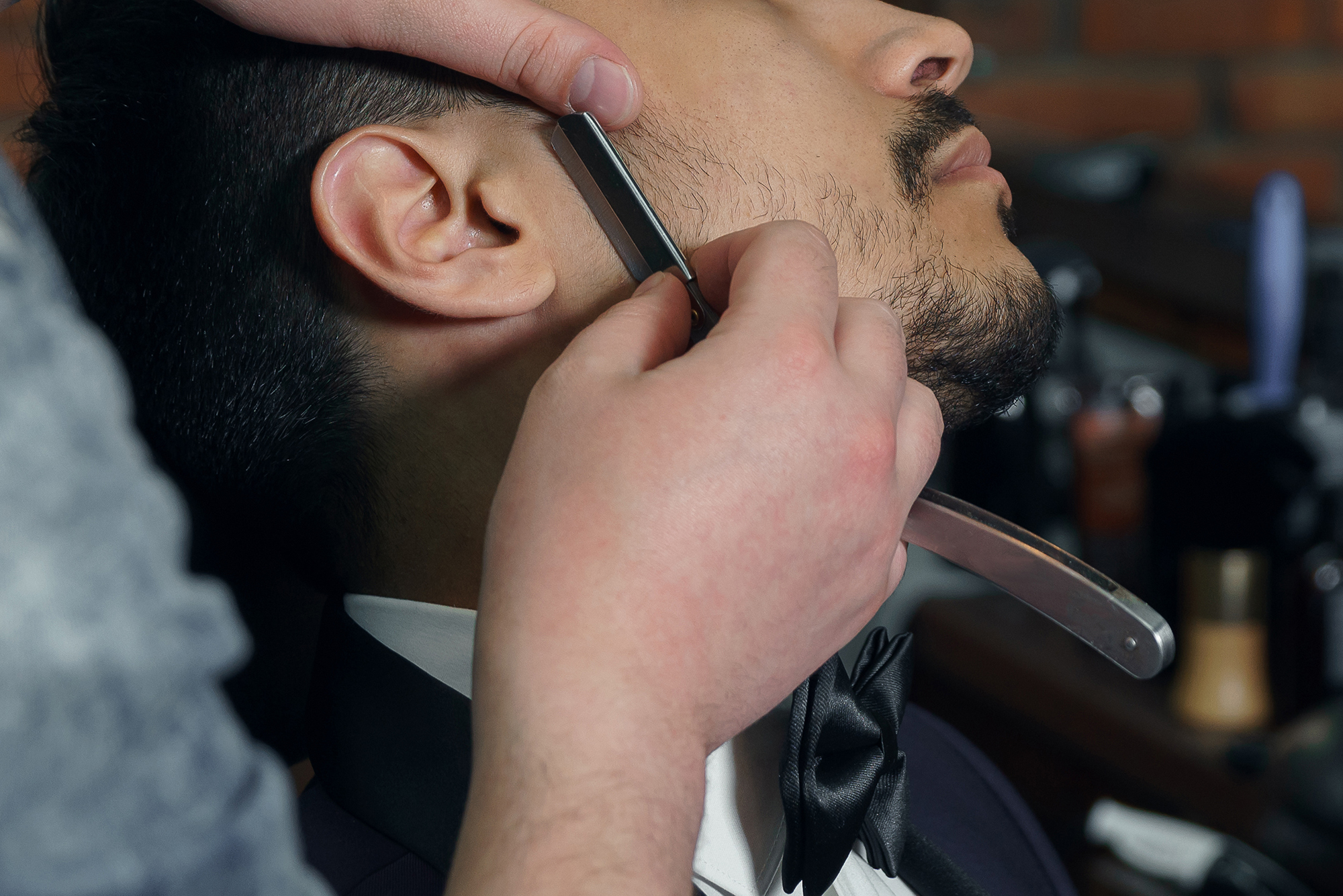Side view of young bearded man in suit getting beard haircut at hairdresser while sitting in chair at barbershop